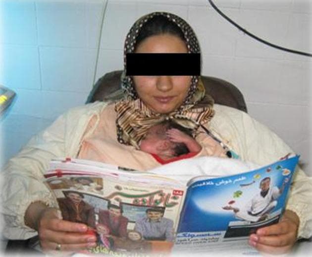 This clinical photograph illustrates Kangaroo Mother Care (KMC) being performed in a hospital setting, likely a Neonatal Intensive Care Unit (NICU). The image shows a mother seated in a supportive chair, holding a premature newborn in a skin-to-skin, vertical position against her chest. The infant is securely nestled within a specialized medical garment, often referred to as a 'Kanga Carrier' or 'Kangaroo Bag,' designed to provide support while keeping the mother's arms free. The newborn appears small with visible reddish skin tone and is positioned for optimal physiological stability. The mother is wearing a patterned headscarf and a light-colored top, demonstrating the implementation of KMC while engaged in a relaxing activity (reading). This visual highlights a key neonatology practice used to improve thermoregulation, facilitate breastfeeding, and enhance bonding for preterm infants. The setting emphasizes the clinical relevance of KMC as a routine, cost-effective intervention for high-risk neonates with stable respiratory and cardiovascular status.
