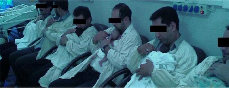 A clinical photograph depicting a row of adult males performing Kangaroo Mother Care (KMC), also known as skin-to-skin contact, within a Neonatal Intensive Care Unit (NICU). The men are seated in specialized chairs, wearing hospital-provided gowns designed to facilitate direct contact with newborn infants held against their chests. The newborns are positioned in a vertical or semi-vertical orientation. In the background, hospital infrastructure is visible, including neonatal cribs and wall-mounted electrical panels with medical-grade outlets. This image illustrates the involvement of fathers in developmental supportive care for preterm or low-birth-weight infants. The educational focus is on non-pharmacological interventions to stabilize neonatal physiology, promote bonding, and improve clinical outcomes in a NICU setting. Key concepts include neonatal nursing, paternal involvement in pediatrics, and the implementation of skin-to-skin contact protocols.