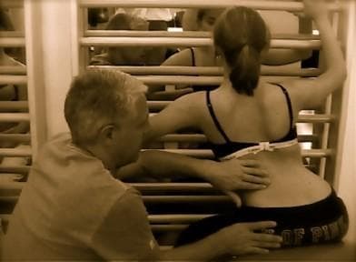 A clinical photograph in a physical therapy setting demonstrating the 'New Power Schroth' exercise for scoliosis management. The image shows a female patient in a kneeling or squatting position facing a wall bar apparatus. Her right arm is elevated and gripping a high bar to facilitate thoracic derotation and redression of a rib hump on the convex side of the spinal curve. A therapist is positioned behind the patient, using tactile cues with his hands on her lumbar region and lower back to monitor postural synergy and alignment. The patient is wearing a dark sports bra with a white measuring tape or band secured around the lower rib cage/waistline to monitor thoracic expansion or spinal correction. This illustration highlights the application of the Schroth method, a specialized physiotherapy approach used to improve pulmonary function, reduce pain, and halt curve progression in adolescent or adult idiopathic scoliosis through three-dimensional spinal correction.