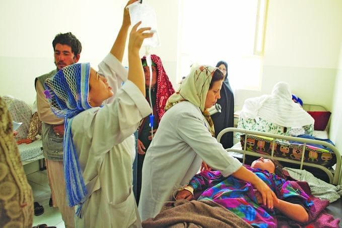 This clinical photograph depicts a healthcare setting in a resource-limited environment, specifically documenting the administration of intravenous (IV) therapy for an obstetric emergency. A female patient, identified by the context as having pre-eclampsia, is lying supine in a hospital bed. A healthcare professional in a white clinical coat is actively managing the IV setup, holding a clear plastic infusion bag (containing magnesium sulphate) elevated above the patient to facilitate gravity-fed flow. The IV tubing is visible, extending from the bag to an insertion point on the patient's arm. The image illustrates critical maternal health interventions and the clinical management of hypertensive disorders of pregnancy. The surrounding individuals and their attire, including headscarves and traditional garments, provide cultural context for the clinical setting in Afghanistan. This visual serves as an educational resource for global health, emergency obstetric care, and the practical application of magnesium sulphate protocols in preventing eclamptic seizures.