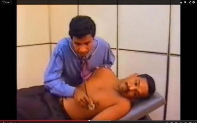 A clinical photograph captured as a video frame depicting a clinician performing a physical examination on a male patient. The clinician is leaning over the patient and using a stethoscope to perform auscultation of the chest. The patient is lying in a supine position on a clinical examination table with the upper body exposed, facilitating access to thoracic landmarks. The clinician is shown positioning the stethoscope diaphragm over the cardiac or pulmonary regions, likely assessing heart sounds (cardiovascular system) or breath sounds (respiratory system). This image illustrates the standard clinical procedure for non-invasive diagnostic monitoring and physical assessment in a medical education context. Relevant medical concepts include auscultation, the cardiovascular examination (CVS), respiratory system (RS) assessment, and the use of medical devices in patient care.