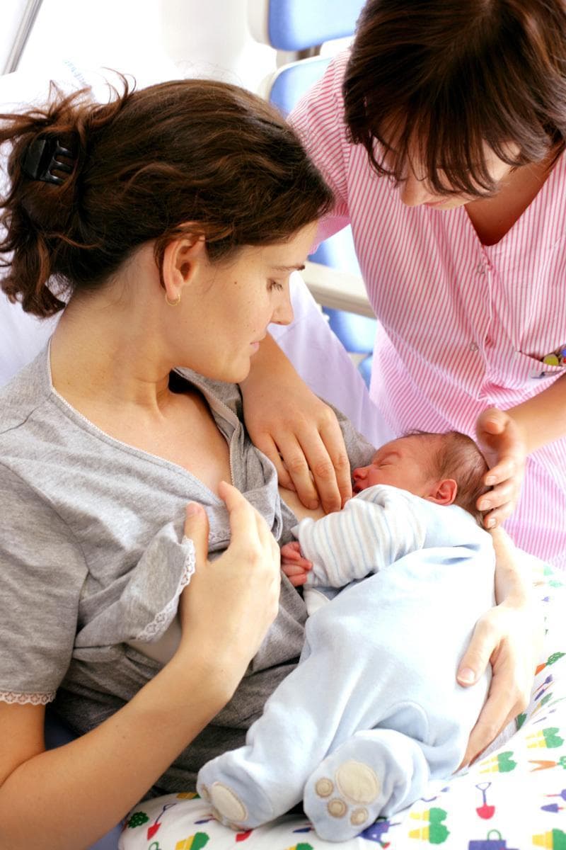 A clinical photograph depicting postpartum lactation support in a healthcare setting. The image shows a mother in a hospital environment seated and breastfeeding a newborn infant. A lactation consultant or nurse, wearing a professional striped uniform, is positioned beside the mother, providing manual guidance to facilitate proper positioning and latch-on technique. The infant is supported by a patterned nursing pillow to optimize ergonomic alignment. This visual illustrates key concepts in neonatal care, maternal education, and the clinical management of breastfeeding. The background includes elements of a hospital bed, identifying the setting as a maternity ward or obstetric unit. This content is designed for medical education focusing on maternal-child health, nursing interventions, and the mechanics of early breastfeeding support.
