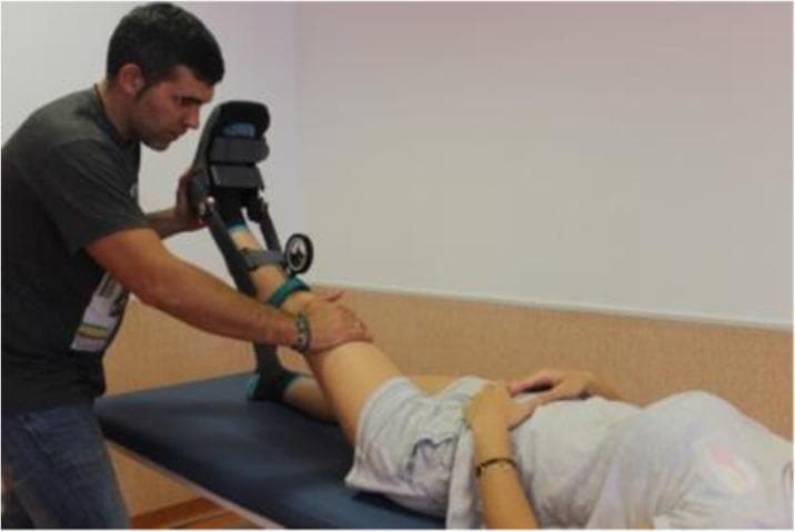A clinical photograph demonstrating a modified Straight Leg Raise (SLR) test, a common physical examination maneuver used to assess lumbar nerve root irritation or hamstring flexibility. The image shows a patient in a supine position on a medical examination table with their hands resting on their abdomen. A clinician stands at the patient's left side, performing passive hip flexion with the knee extended. The patient's left leg is secured within a specialized mechanical orthosis or goniometric device, which is fastened with green adjustable straps and features an integrated circular dial or inclinometer to quantify the angle of elevation. The clinician's right hand is placed superior to the patient's patella to maintain knee extension during the procedure. This procedural image illustrates the clinical application of objective measurement tools in musculoskeletal assessment and physical therapy, focusing on diagnostic accuracy for conditions such as lumbar disc herniation or sciatica.