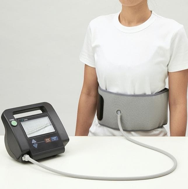 A clinical photograph of an innovative medical exercise device designed for abdominal trunk muscle strengthening and strength measurement. The setup consists of a wide, gray inflatable fabric cuff positioned around the subject's midsection and a black mechanical manometer resting on a flat surface. A flexible gray connecting tube links the cuff's anterior port to the manometer's base. The manometer features a digital screen displaying real-time pressure waveforms (measured in kPa), alongside a power button and operational controls. This device is utilized in physical therapy and rehabilitation settings to facilitate lumbar stabilization exercises and measure core muscle strength through intermittent abdominal contractions against baseline pressure. The system is particularly relevant for managing chronic lower back pain and improving spinal stability by activating the diaphragm, abdominal, and pelvic floor muscles without requiring painful spinal flexion or extension.