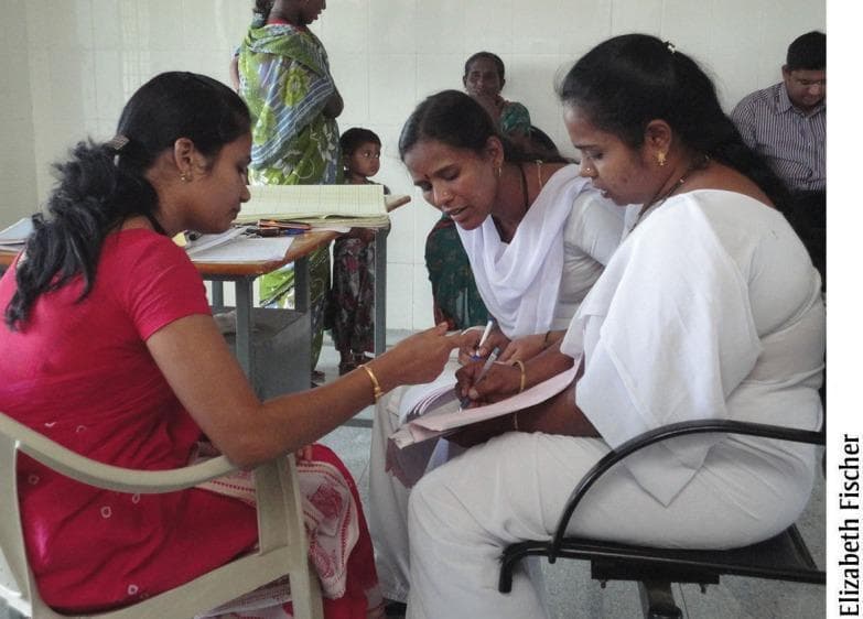 A clinical photograph depicting a healthcare training session in a primary health center in Karnataka, India. The image shows a nurse mentor, dressed in a red top and patterned garment, providing clinical support and instruction to two staff nurses. The staff nurses are wearing traditional white uniforms with white scarves. They are engaged in a case sheet audit, with one nurse actively writing on a patient medical record while the mentor points to specific documentation sections. This interaction illustrates quality improvement processes and provider skill upgrading through bedside case reviews and modeling of good practices. In the background, other individuals, including a child and a woman, are visible in the clinical environment. The photograph serves as an educational example of maternal and neonatal health mentoring interventions in rural, low-resource settings, focusing on improving the accuracy of clinical documentation and the quality of institutional delivery care.
