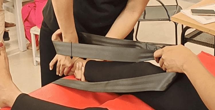 This clinical photograph demonstrates a physical therapy manual mobilization technique for the ankle joint. A therapist is performing a posterior talar glide in a non-weight-bearing position to increase the range of motion for ankle dorsiflexion. The patient is positioned supine on a treatment table. The therapist utilizes a stabilization strap (Mulligan-style belt) looped around their own waist and the patient’s distal tibia/fibula to provide stabilization and traction. The therapist's hands are placed specifically over the anterior aspect of the talus to apply a posterior force, indicated by a black directional arrow. This orthopedic intervention is commonly used in the management of joint stiffness related to conditions like plantar fasciitis, heel spurs, or post-immobilization stiffness. The image illustrates the clinical application of mobilization with movement (MWM) principles within sports medicine and musculoskeletal rehabilitation.