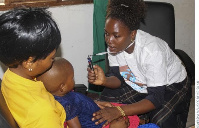 This clinical photograph illustrates a pediatric ophthalmic examination using the Arclight, a low-cost, portable, solar-powered direct ophthalmoscope and otoscope. The image shows a healthcare professional in a clinical setting in Tanzania performing an eye screening on a young child who is being held by a caregiver. The examiner is holding the slender, black, handheld device in her right hand, positioned at a close working distance to the child's eye to assess the red reflex or perform a fundoscopic examination. The device features a white neck lanyard for easy accessibility during field screenings. This visual demonstrates the practical application of frugal innovation in global health settings for the early detection of ocular conditions such as cataracts, retinoblastoma, and refractive errors in pediatric populations. The clinical focus is on the technique of direct ophthalmoscopy and the use of specialized diagnostic tools adapted for resource-limited environments.