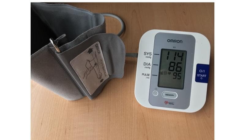 A clinical photograph of a portable electronic sphygmomanometer, specifically an Omron M3 digital blood pressure monitor, used for cardiovascular health assessment. The image displays the digital base unit and the associated upper-arm cuff. The LCD screen on the base unit shows live readings: Systolic (SYS) blood pressure at 114 mmHg, Diastolic (DIA) blood pressure at 86 mmHg, and a pulse rate of 95 beats per minute (bpm). To the left, the gray fabric blood pressure cuff is visible, featuring a metal D-ring for secure tightening and a printed instructional label. This label provides a visual diagram demonstrating correct cuff placement relative to the elbow joint and brachial artery to ensure measurement accuracy. The device is a standard tool for monitoring hypertension and heart rate in clinical and home settings. Key educational components include the interface of digital diagnostic equipment and the importance of standardized placement for reliable vital sign data collection.