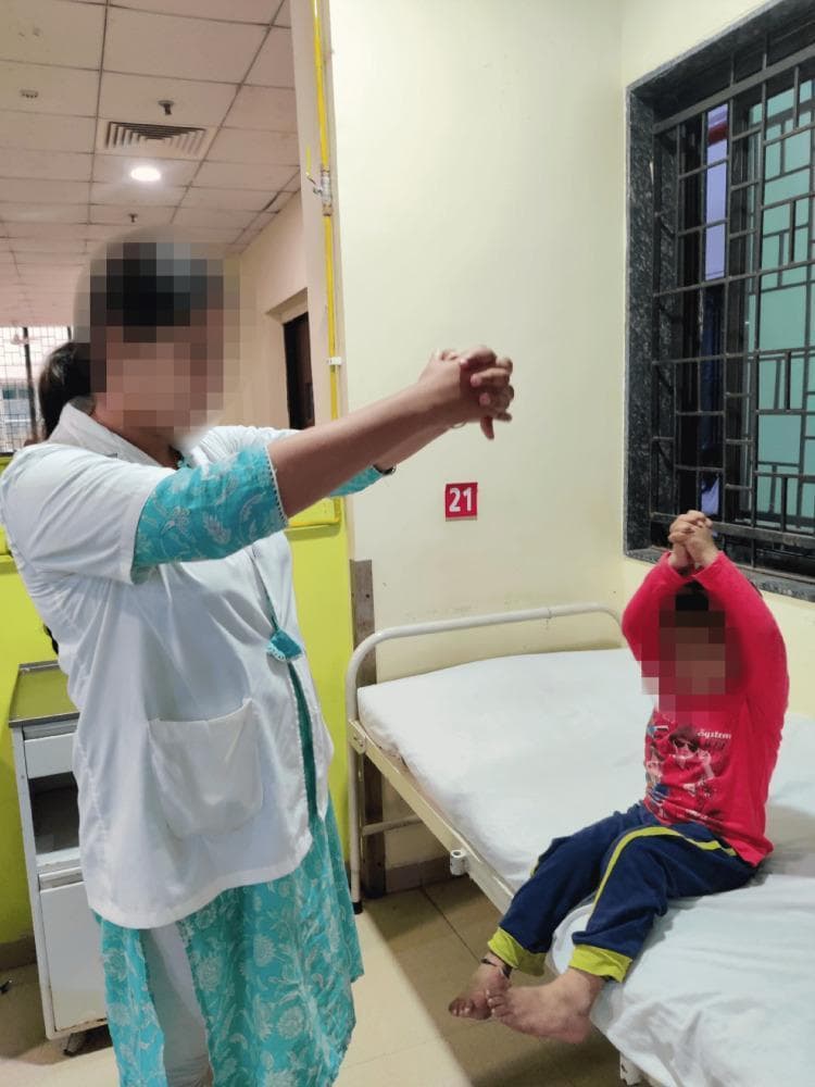 This clinical photograph captures a pediatric patient and a healthcare professional engaged in a respiratory physiotherapy session within a hospital setting. The patient, a young child, is shown in a long-sitting position on a hospital bed with legs extended. Guided by the therapist, the child is performing thoracic expansion exercises, characterized by the raising of both arms overhead with hands clasped to facilitate maximum lung volume and chest wall mobility. The healthcare professional, wearing a white clinical coat, stands adjacent to the bed and demonstrates the movement with their own arms extended and hands clasped. This intervention is typical for post-operative cardiac rehabilitation (following median sternotomy for VSD repair, as indicated by context) to prevent atelectasis and improve pulmonary function. The background reveals a standard clinical environment with a numbered patient station and safety-barred window. This image serves as an educational example of active-assisted breathing techniques and the role of physical therapy in pediatric cardiac recovery.