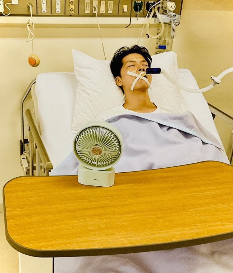 Clinical photograph of a patient in an intensive care unit (ICU) setting demonstrating the application of 'fan therapy' for dyspnea management. The patient is supine in a hospital bed, appearing sedated or with eyes closed, and is undergoing mechanical ventilation via an endotracheal tube secured with adhesive tape. In the foreground, a portable, battery-operated electric fan is positioned on an overbed tray table, directed toward the patient's face. This non-pharmacological intervention is designed to alleviate the sensation of breathlessness by providing cool airflow to the regions innervated by the second (maxillary) and third (mandibular) branches of the trigeminal nerve. The background shows standard critical care infrastructure, including medical gas outlets, electrical panels, and suction equipment. This image illustrates palliative and supportive care techniques used to manage respiratory distress in critically ill or end-of-life patients.