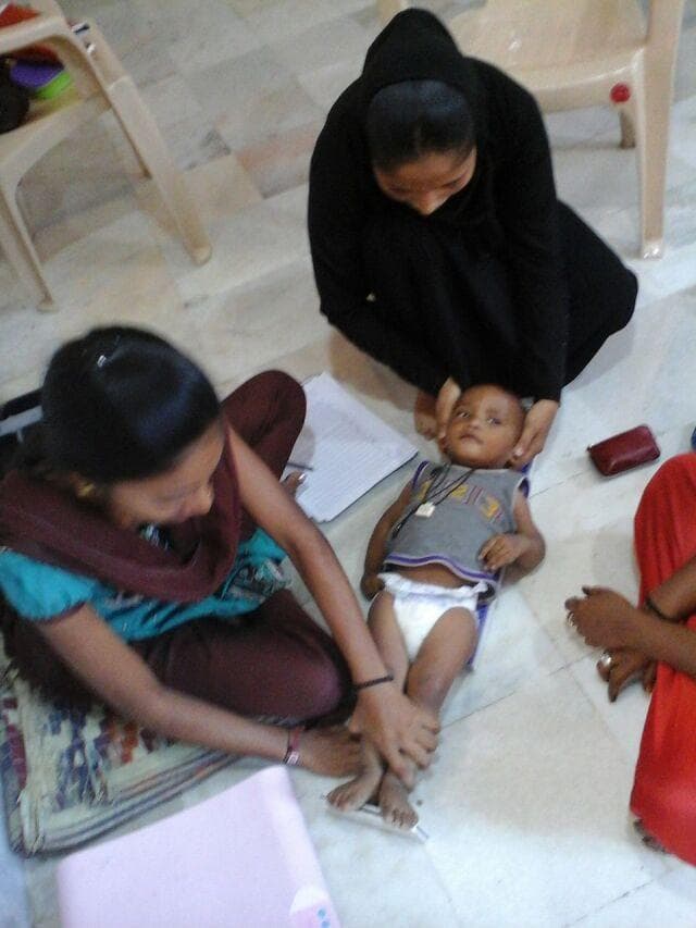 A clinical photograph depicting growth monitoring and anthropometric assessment of an infant by frontline health workers in a community health setting. The infant is positioned supine on a floor surface for length measurement. One health worker is situated at the head, ensuring proper alignment and stabilization of the infant's crown against a headboard. A second health worker is positioned at the infant's feet, extending the legs and ensuring the feet are flexed against a footboard to obtain an accurate recumbent length measurement. Essential medical equipment and documentation tools are visible, including a pink infant weighing scale, a notebook for data recording, and a pen. This image illustrates the standardized procedures for screening and monitoring pediatric nutritional status, specifically as part of a Community-Based Management of Acute Malnutrition (CMAM) program. The educational focus is on the clinical technique for infant anthropometry and the role of community-based health interventions in pediatric primary care.