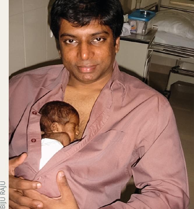 A clinical photograph illustrating Kangaroo Mother Care (KMC) performed by a male caregiver, demonstrating the practice of skin-to-skin contact for a preterm infant. The photograph shows an adult male of South Asian descent holding a small neonate against his bare chest, tucked inside a partially unbuttoned shirt to maintain warmth and bonding. The infant appears to be a premature newborn with dark hair, resting in a vertical position. This image serves as an educational example of neonatal supportive care, particularly relevant in the management of preterm births and the prevention of complications such as Retinopathy of Prematurity (ROP) through physiological stabilization. The setting appears to be a neonatal clinical environment, emphasizing the role of paternal involvement in evidence-based neonatal interventions in resource-limited or high-burden settings like India.