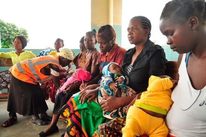 A clinical photograph depicting a maternal and child health (MCH) outreach setting at the Lundazi Urban Health Centre in Zambia. The image illustrates a group of mothers and their infants seated on a wooden bench within a basic clinical facility, characterized by painted walls and concrete flooring. A member of the Safe Motherhood Action Group (SMAG), identified by a high-visibility orange safety vest, is shown interacting with an infant held by one of the seated women. This visual documents community-level health systems strengthening and the 'Continuum of Care' (CoC) model designed to improve access to basic and emergency obstetric and newborn care. The primary educational focus is on global health initiatives, specifically the Saving Mothers, Giving Life (SMGL) project, which utilizes community health workers and peer supporters to bridge the gap between rural populations and skilled birth attendants. This photograph serves as a real-world example of demand-side health interventions and the mobilization of community health volunteers in maternal, newborn, and child health (MNCH) programs in sub-Saharan Africa.