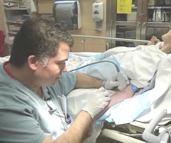 Clinical photograph of a procedural intervention in an emergency department or clinical setting, demonstrating ultrasound-guided intravenous (IV) catheter placement. A clinician, wearing non-sterile medical gloves, is shown performing the procedure on a patient's antecubital region. The image illustrates the long-axis (longitudinal) approach, where a high-frequency linear array ultrasound probe is held parallel to the course of the target brachial vein to visualize the needle's trajectory and vessel entry in real-time. A blue tourniquet is applied to the patient's upper arm to facilitate venous distension. The environment includes standard medical infrastructure, such as a hospital bed, blue drapes, and bedside supplies. This procedural image highlights clinical best practices for difficult vascular access, emphasizing the use of point-of-care ultrasound (POCUS) to improve first-attempt success rates and reduce complications associated with peripheral cannulation in the antecubital fossa.