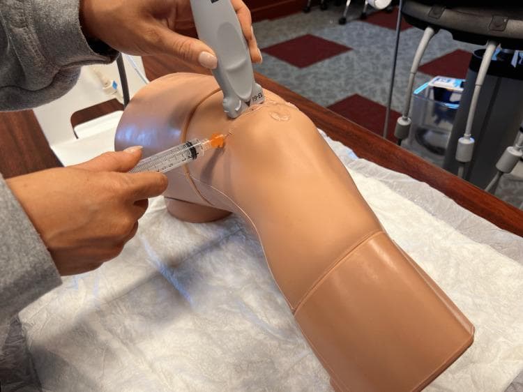 This procedural photograph illustrates a demonstration of an ultrasound-guided knee aspiration (arthrocentesis) on a medical simulator. The image shows a realistic, light-brown anatomical model of a human knee resting on a white clinical drape. A practitioner's hand is seen holding a gray linear ultrasound transducer positioned in the superolateral aspect of the patella, with clear conductive gel visible at the contact point. Simultaneously, another hand is inserting a clear plastic syringe equipped with a fine-gauge needle (indicated by the orange hub) into the joint space. The needle is oriented parallel to the transducer's long axis, demonstrating the 'in-plane' needle visualization technique common in musculoskeletal procedures. This setup teaches medical students how to utilize real-time imaging to guide needle placement into the suprapatellar bursa for diagnostic fluid aspiration or therapeutic injection. The context emphasizes procedural accuracy, landmarking, and the integration of ultrasound technology in clinical skills training for orthopedic or emergency medicine specialties.