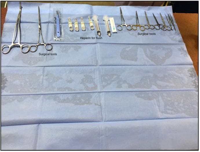This intraoperative clinical photograph displays a sterile-draped workbench organized for surgical organ retrieval, specifically in an experimental or translational research setting. The instruments are laid out in a linear, structured arrangement on a blue sterile field. On the far left, two large long-reach curved hemostatic clamps (forceps) are placed, used for heavy tissue manipulation or vessel clamping. To their right, a sterile-packaged scalpel and four glass ampules of heparin are visible, the latter designated for preparing heparinized saline flushes to prevent thrombosis in harvested vasculature. Adjacent to these are two plastic syringes and a hypodermic needle. On the far right, a variety of stainless steel precision instruments are organized by size, including multiple pairs of surgical scissors, finer hemostats, and anatomical forceps (tweezers) for delicate dissection of small vessels and ligaments. The setup demonstrates the standard clinical requirements for identifying and resecting key anatomical structures such as the hepatic artery and portal vein during organ isolation.