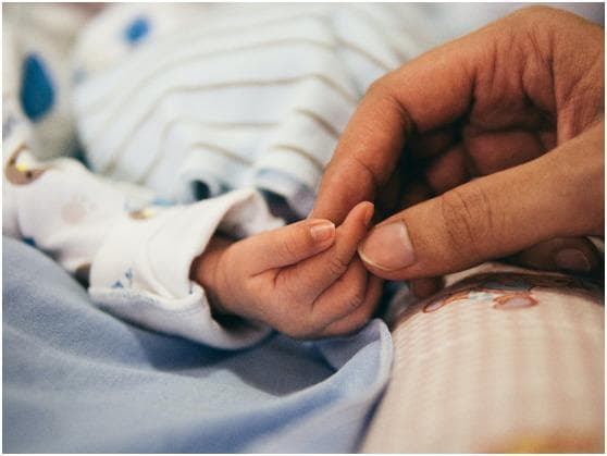 A high-detail clinical photograph capturing the physical interaction between an adult and a neonate, illustrating relative scale and developmental anatomy. The image focuses on the hands, where an adult's index finger and thumb are gently grasping the smaller hand of an infant. This comparison highlights the significant difference in hand size, skin texture, and nail morphology between developmental stages. The infant is shown in a clinical or home care setting, wearing a patterned long-sleeved garment and resting on blue and pink check-patterned bedding. The image demonstrates the palmar grasp reflex or general neonatal bonding, serving as a visual reference for pediatric physical examination, neonatal development, and maternal-infant health education. The lighting is soft and natural, emphasizing the fine skin folds of the infant's hand against the larger, mature hand of the adult.