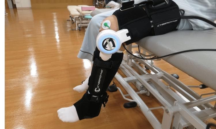 A clinical photograph depicting a patient undergoing postoperative rehabilitation following a Total Knee Arthroplasty (TKA). The image shows a single-joint Hybrid Assistive Limb (HAL-SJ) robotic exoskeleton attached to the patient's lower extremity. The device is secured to the thigh and calf with black padded 'CYBERDYNE' straps, with its central motor unit aligned precisely over the lateral aspect of the knee joint. Surface electromyography (sEMG) electrodes are visible on the distal vastus medialis, utilized to detect bio-electrical signals (BES) for cybernic voluntary control. The patient is positioned on a medical treatment table in a rehabilitation gym setting, performing knee extension and flexion exercises. This robotic intervention is used in physical therapy to assist active-assisted mobilization and neuromuscular gait re-education by providing torque support triggered by the patient's muscle signals. The photograph illustrates a procedural application of advanced medical robotics in orthopedic recovery for conditions like knee osteoarthritis.