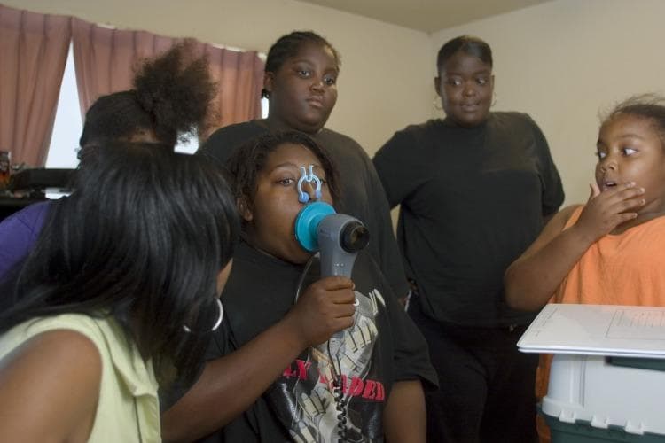 A clinical photograph depicting a pediatric patient using a handheld peak flow monitor (spirometry device) in a community or home setting. The central figure is a young girl performing a pulmonary function test; she has a blue circular mouthpiece sealed over her lips and a light blue nose clip applied to prevent nasal airflow. She holds the gray cylindrical handle of the device, which is connected by a coiled black cable to an unseen recording base. Surrounding her are other children observing the procedure. This image illustrates the practical application of respiratory diagnostic tools in community health or clinical research settings, specifically focusing on asthma management and the assessment of Peak Expiratory Flow (PEF). The clinical significance relates to monitoring airway obstruction and lung capacity in pediatric populations.