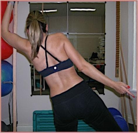 Clinical photograph from a posterior view showing a female patient with idiopathic scoliosis performing a Schroth method physiotherapy exercise known as the 'muscle cylinder.' The image captures visible postural deviations, including a right-sided thoracic rib hump, an elevated right shoulder, and a depressed, retracted left shoulder. The patient is engaged in active axial self-elongation and 3D postural correction. Her left arm is elevated and reaching overhead to facilitate expansion of the thoracic concavity and spinal de-rotation, while her right arm is extended laterally for stabilization. This therapeutic intervention aims to address a 3C curve type (according to Schroth classification) by utilizing rotational breathing techniques to increase vital capacity and reduce the Cobb angle. The setting is a rehabilitation environment with physiotherapy equipment, such as stall bars and exercise balls, visible in the background, illustrating the practical application of conservative scoliosis management.