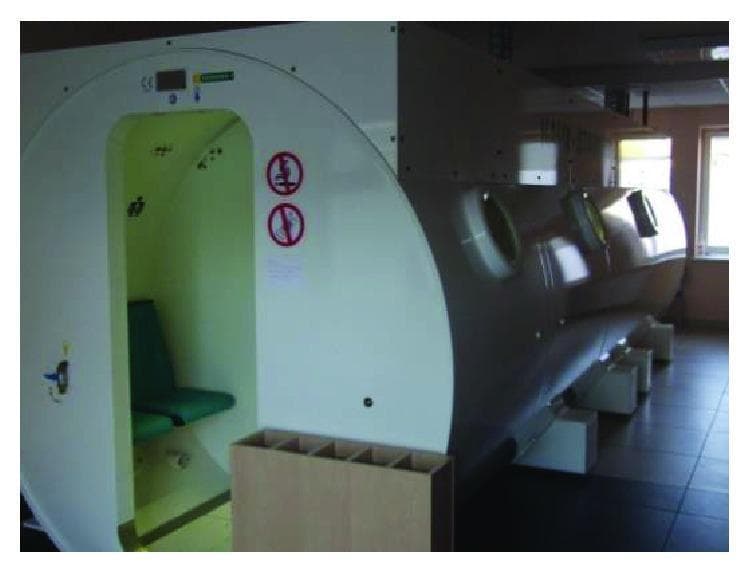 Clinical photograph of a multiplace hyperbaric oxygen therapy (HBOT) chamber, identified as a Haux Starmed 2200 model, located within a specialized medical facility. The chamber is a large, cylindrical, pressure-sealed vessel designed for delivering oxygen at pressures higher than atmospheric levels. The exterior features a dual-color scheme with a white front-facing entrance panel and a grey longitudinal hull. The entrance consists of a reinforced doorway revealing an illuminated interior with green patient seating. Circular, thick-paned pressure windows (portholes) are distributed along the side for patient monitoring. External safety signage, including red 'prohibition' symbols (likely for fire hazards or smoking), is visible near the entry point. The unit is supported by heavy-duty white rectangular structural blocks on a tiled clinical floor. Such devices are used in the management of conditions like Sudden Sensorineural Hearing Loss (SSNHL), chronic non-healing wounds, and decompression sickness, where hyperbaric oxygen serves to increase plasma-dissolved oxygen and modulate oxidative stress markers.