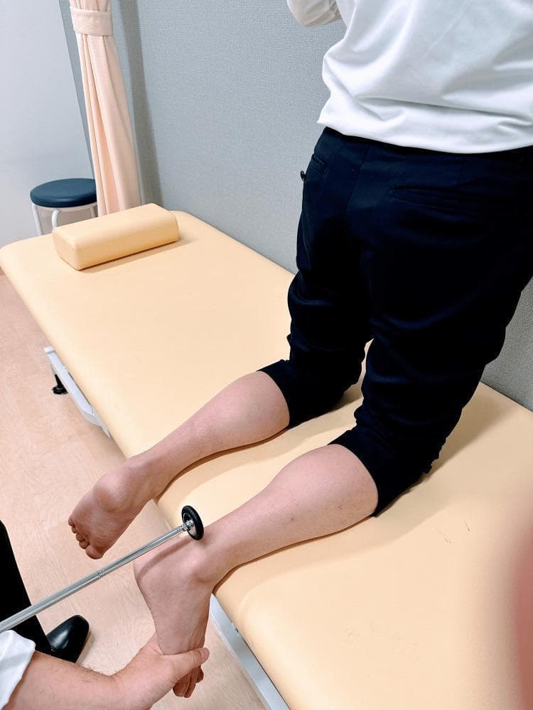 A clinical photograph demonstrating the kneeling technique for eliciting the Achilles tendon reflex (ankle jerk), which tests the S1-S2 nerve roots and the tibial nerve. The patient is positioned kneeling on an examination table with their shins supported by the table surface and their feet hanging freely over the edge. This position allows for optimal relaxation of the gastrocnemius-soleus complex. The examiner is shown performing two simultaneous actions: using one hand to apply gentle passive dorsiflexion to the plantar surface of the foot to place the Achilles tendon under slight tension, and using a standard reflex hammer in the other hand to strike the tendon just above its insertion on the calcaneus. This procedural image illustrates a standard method in a neurological physical examination to assess deep tendon reflexes and identify possible upper or lower motor neuron pathology.