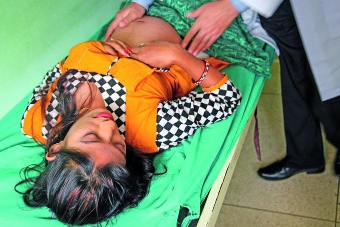 This clinical photograph captures an obstetric physical examination in a community or public health setting. A female patient is lying supine on an examination table with her abdomen exposed. The gravid abdomen is clearly visible, demonstrating significant distension consistent with pregnancy. The hands of a healthcare provider are positioned on the uterine fundus and lateral aspects of the abdomen, performing Leopold's maneuvers or a similar abdominal palpation to assess fetal position, presentation, or fundal height. The patient is wearing colorful traditional clothing, and the clinician is dressed in professional attire, including a white lab coat. The image illustrates a core procedural skill in maternal healthcare and prenatal screening, highlighting the clinical assessment of gestation within the obstetric and gynecological specialty. It serves as an educational resource for demonstrating patient positioning and manual examination techniques in low-resource or community health contexts.