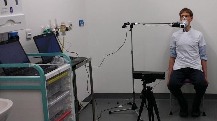 This clinical photograph illustrates a dual-modality respiratory assessment setup designed to correlate traditional spirometry with 3D motion tracking. A male subject is seated upright wearing a tight-fitting white t-shirt, which serves to minimize loose fabric and optimize the accuracy of chest wall surface tracking. He is positioned facing a Microsoft Kinect sensor mounted on a tripod at approximately chest height, used to capture thoracic wall volume changes and respiratory rate via depth-sensing technology. Simultaneously, the subject is connected to an unmodified digital spirometer through a mouthpiece and nose clip to measure standard pulmonary function parameters, including Forced Vital Capacity (FVC) and Forced Expiratory Volume (FEV1). The spirometer tubing is supported by a microphone stand to prevent the equipment from resting on the chest or obscuring the camera's view. In the background, two laptops on a medical cart facilitate real-time data acquisition and temporal alignment of the volume-time curves from both devices. This setup is typical for clinical research into non-contact respiratory monitoring and the validation of optoelectronic plethysmography techniques.