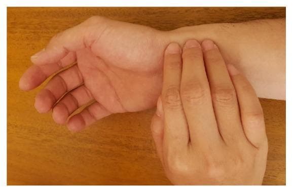 This clinical photograph demonstrates the manual technique for pulse diagnosis, specifically within the context of Traditional Chinese Medicine (TCM). The image shows a patient's right forearm and wrist in a supinated position (palm facing up) resting on a wooden surface. A practitioner is performing palpation using three fingers—the index, middle, and ring fingers—placed along the radial artery at the distal end of the radius. The fingers are aligned longitudinally from the wrist crease toward the elbow, corresponding to the TCM anatomical positions known as the 'inch' (Cun), 'bar' (Guan), and 'cubit' (Chi). The practitioner's fingertips are slightly curved and apply gentle pressure to the radial pulse to assess vascular qualities. This visual illustrates the traditional 'three positions and nine indicators' method used to evaluate organ health and systemic balance through arterial pulse characteristics. The photograph highlights the specific anatomical placement required for accurate manual arterial assessment in a clinical or educational setting.