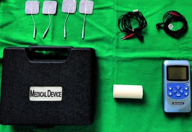 Clinical photograph of a transcutaneous electrical nerve stimulation (TENS) device kit, commonly used in physical therapy, pain management, and experimental medicine—such as parotid gland stimulation to increase salivary flow. The components are arranged on a green sterile field and include a dual-channel TENS control unit (model MH8000), four square self-adhesive electrode pads with integrated lead wires, two coiled black electrical cables with dual-terminal connectors, a roll of micropore medical tape for securing electrodes, and a black hard-shell carrying case labeled 'MEDICAL DEVICE'. The blue control unit features a digital LCD screen and tactile interface buttons for adjusting pulse rate, pulse width, and intensity (0-80mA). This visual serves to illustrate the equipment required for non-invasive neurostimulation procedures in both clinical and research settings.