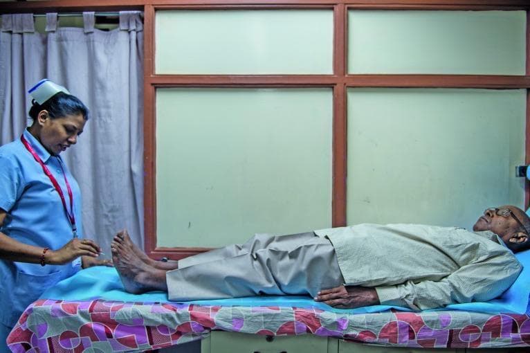 A clinical photograph capturing a diabetic foot examination in a healthcare setting. The image shows a medical professional, identified as a nurse in a blue scrub suit and cap, performing a sensory assessment on an elderly male patient. The patient is lying supine on an examination table. The nurse is utilizing a specialized instrument, consistent with a Semmes-Weinstein monofilament, to test for peripheral neuropathy by applying pressure to specific points on the patient's toes and plantar surface. This procedure is a critical diagnostic step in identifying loss of protective sensation (LOPS) in patients with diabetes mellitus, aimed at preventing ulcers and complications related to diabetic foot disease. The setting includes clinical furniture, patterned linens, and privacy curtains, illustrating a standard outpatient diabetic screening environment.