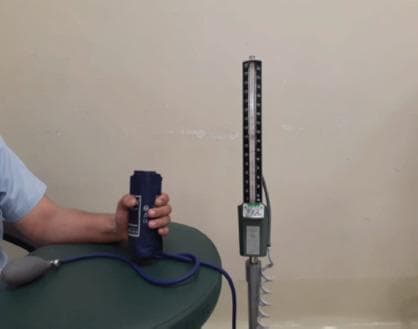 Clinical photograph of a patient's hand being used to assess hand grip strength using a modified mercury sphygmomanometer. The setup includes a dark blue blood pressure cuff that has been rolled into a cylindrical shape to fit the patient's palm, connected via blue flexible tubing to a hand-held inflation bulb and a vertical mercury-filled column scale. The scale is mounted on a metallic base and shows numerical gradations for pressure measurement in mmHg. This clinical assessment technique is often utilized in postoperative hand rehabilitation, such as after ulnar claw hand deformity correction, to provide a reproducible and reliable measurement of functional grip strength. The photograph captures the educational application of standard diagnostic equipment for monitoring recovery in orthopedic and plastic surgery contexts.