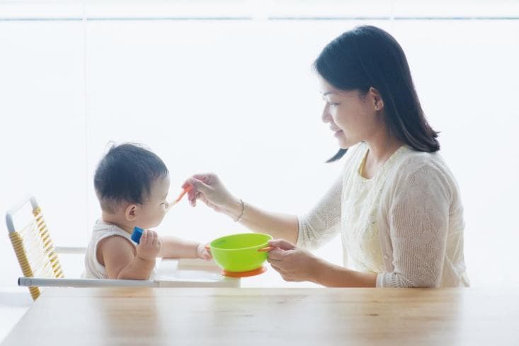 This clinical photograph illustrates the process of complementary feeding in an infant, typically occurring between 5 to 12 months of age. The image shows a caregiver (mother) introducing semi-solid food to an infant seated in a high chair. The mother is using a small, orange plastic spoon to deliver food from a green and orange bowl, demonstrating the transition from exclusive liquid nutrition (breastmilk or formula) to solid foods. The infant is actively engaged, holding a small object in one hand, which reflects normal developmental motor skills associated with this age group. This stage is medically significant as a critical window for establishing lifelong dietary patterns, promoting growth, and diversifying the gut microbiome. The visual emphasizes the manual coordination required during the weaning period and the role of caregiver-led feeding in ensuring adequate nutrient intake, particularly protein and fiber, to prevent undesired growth patterns or obesity later in life.