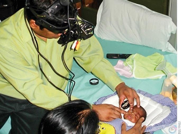 A clinical photograph depicting a pediatric ophthalmic examination and treatment session in a medical facility. An ophthalmologist is seen wearing a specialized head-mounted indirect ophthalmoscope or laser delivery system, equipped with an integrated light source and visible AV/data cables. The clinician is using a handheld condensing lens to perform an eye examination on an infant, who is lying on a padded surface and being stabilized by an assistant. The clinical context suggests a screening or treatment evaluation for Retinopathy of Prematurity (ROP), potentially involving peripheral retinal photocoagulation. The scene illustrates the technical setup required for neonatal ophthalmology, including the use of magnification, focused illumination, and specialized pediatric positioning. Key educational elements include the demonstration of indirect ophthalmoscopy technique and the management of neonatal retinal vascular diseases.