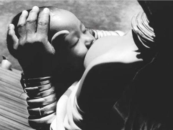 A black-and-white clinical photograph documenting an infant breastfeeding, serving as an educational example of maternal-child health and nutrition in global health settings. The image illustrates the 'cradle hold' positioning, with the infant's face turned toward the breast and a deep latch visible. The adult provides manual support to the infant's occiput to facilitate stable positioning during feeding. Notable cultural markers include stacked brass neck rings and wrist coils on the adult, which are characteristic of specific ethnic groups in Southeast Asia. This visual emphasizes the clinical significance of breastfeeding as a critical intervention for infant survival and immune support, particularly in resource-limited or emergency environments like refugee camps. Key educational concepts include lactation management, infant latch technique, and the sociocultural context of neonatal care.