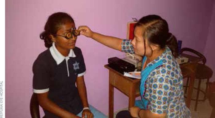 This clinical photograph captures a subjective refraction procedure being performed in a community eye care setting. A seated adolescent patient is wearing a metallic universal trial frame, which is fitted with multiple corrective lenses. An ophthalmic assistant is shown manually adjusting the trial frame to ensure proper centration and vertex distance. The procedure is a critical step in diagnosing refractive errors such as myopia, hyperopia, or astigmatism and determining a patient's final spectacle prescription. The scene demonstrates a typical outreach or school-based vision screening environment, highlighting the use of trial frames and lens sets as essential tools for refractive assessment in public health ophthalmology. Educational focus includes proper technique for fitting trial frames, the patient-provider interaction during vision testing, and the clinical application of optometric instruments in low-resource or community-based medical settings.