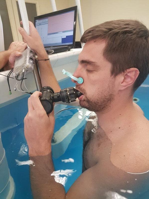 This clinical photograph illustrates a research setup for measuring static respiratory compliance during head-out-of-water immersion. A male subject is shown immersed in water up to the level of the sternal notch, a condition that physiologically alters respiratory mechanics due to hydrostatic pressure on the chest wall. The subject is wearing a nose clip to ensure a closed respiratory system, breathing exclusively through a mouthpiece. The mouthpiece is integrated with a pressure sensor and connected via tubing to a calibrated syringe (partially visible) used to manipulate lung volumes. In the background, a computer monitor displays real-time physiological data and waveforms, likely recording the relationship between pressure and volume changes. This experimental design is used in sports medicine and pulmonary physiology to study the effects of immersion on the respiratory system, specifically evaluating parameters like tidal volume, expiratory reserve volume, and overall respiratory system compliance (Crs).