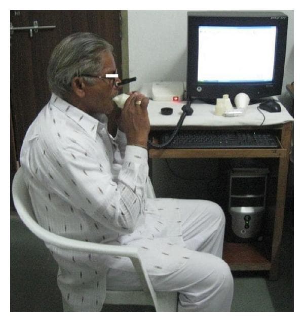 A clinical photograph illustrating a patient performing a Pulmonary Function Test (PFT) via spirometry in a clinical or research setting. The image depicts an older adult male seated upright, wearing a nose clip to ensure mouth-only breathing. He is actively interacting with a handheld spirometer, holding the disposable mouthpiece with both hands to maintain a tight seal. The device is connected via a data cable to a computer system, which includes a monitor displaying an active interface, likely for real-time visualization of flow-volume loops and spirograms. On the desk, several replacement white cylindrical mouthpieces and the hardware interface are visible. This visual serves as an educational example of the standardized setup for measuring lung volumes and ventilation capacities, such as Forced Vital Capacity (FVC) and Forced Expiratory Volume in 1 second (FEV1), highlighting the necessary equipment and proper patient positioning during the procedure.
