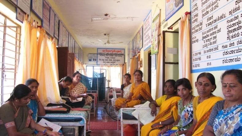This clinical photograph depicts a community healthcare setting in India, representative of primary care delivery and Health Technology Assessment (HTA) implementation at the ground level. The image shows a group of women, dressed in traditional saris, seated within a functional clinical ward or waiting area. The environment is equipped with essential medical furniture, including metal examination beds or cots and a wheelchair visible in the background. The walls are densely populated with educational posters and health charts, many of which utilize local regional scripts (Kannada) for public health communication. This visual illustrates the infrastructure of public health centers in federal healthcare systems, highlighting the interface between medical services and the community. Key educational concepts include global health systems, public health education, and the socio-cultural context of healthcare delivery in low-to-middle-income countries.