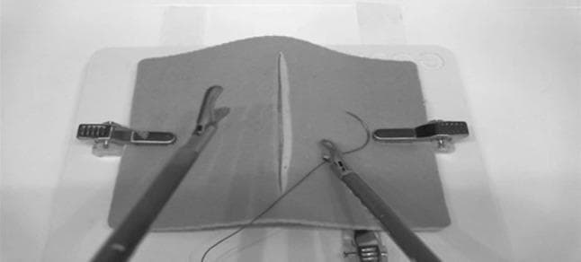 This monochrome image displays a still frame from a laparoscopic surgical simulator, specifically designed for intracorporeal suturing training. The focus is on a central suturing pad with a linear vertical defect that mimics a surgical incision or wound. Two laparoscopic instruments are active within the simulated field: a grasper on the left side and a needle holder on the right. The needle holder is currently manipulating a curved surgical needle attached to a suture thread, demonstrating the initial phase of wound closure. The suturing pad is secured to a white platform by lateral metal clamps. This setup is used in medical education to develop psychomotor skills, depth perception, and manual dexterity required for minimally invasive surgery. The image serves as a clinical simulation tool for teaching advanced surgical techniques, such as continuous or interrupted suturing, within a controlled augmented reality environment.