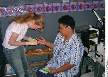 A clinical photograph depicting a volunteer healthcare worker performing a capillary blood glucose test on a patient. The scene takes place within an ophthalmology clinic, evidenced by the presence of a slit-lamp biomicroscope in the background, a tool essential for comprehensive eye examinations. The healthcare worker, wearing protective gloves, is shown preparing a finger-stick puncture on the patient's hand. On a small wooden table, clinical supplies including cotton swabs and lancets are visible. This image illustrates integrated health screening, specifically the importance of monitoring blood sugar levels in patients who may be at risk for diabetic retinopathy or other systemic conditions affecting ocular health. The setting suggests a community health or outreach environment, highlighting patient triage and secondary prevention strategies in specialized medical facilities.