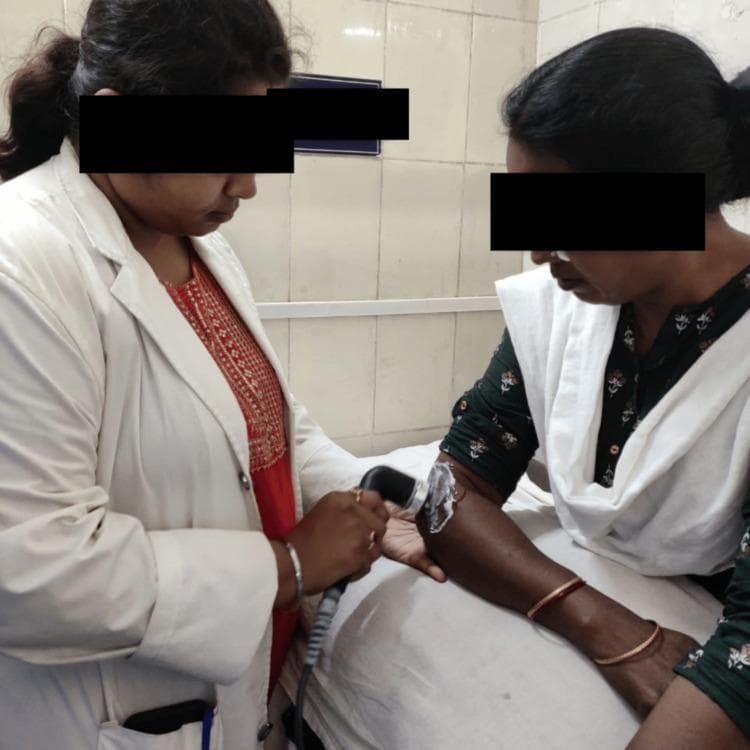 A clinical photograph depicting a physiotherapy session focused on ultrasonic therapy. A female healthcare provider, wearing a white clinical coat, is shown applying a handheld therapeutic ultrasound transducer to the lateral aspect of a female patient's proximal left forearm, near the elbow. The patient's arm is resting on a white examination surface. A visible layer of clear coupling gel is applied to the skin over the treatment site to facilitate the transmission of ultrasonic waves. The ultrasound probe is ergonomic and black, connected via a thick gray cable to a power unit (not shown). This setup is characteristic of conservative management for musculoskeletal conditions such as lateral epicondylitis or soft tissue adhesions, aiming to promote tissue healing and alleviate fibrosis through thermal or non-thermal acoustic energy. The setting is an indoor clinical environment with tiled walls.