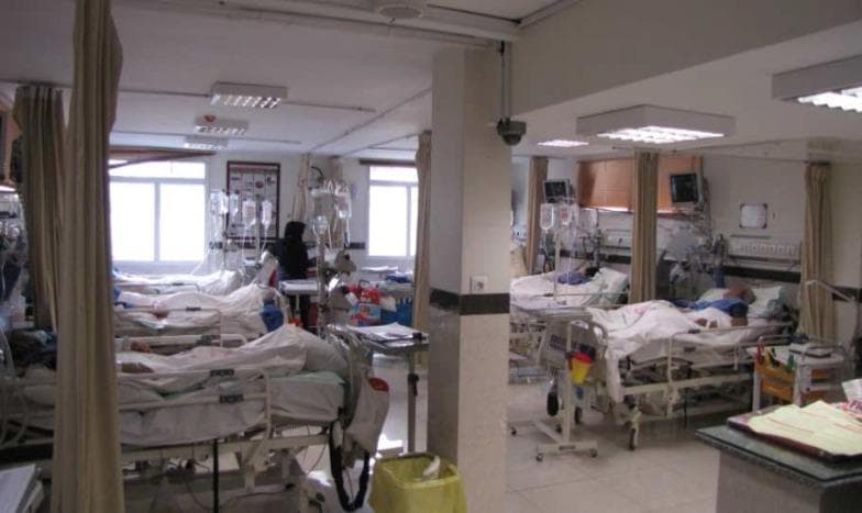 Clinical photograph of a specialized acute stroke unit within a hospital setting. The image demonstrates a multi-bed ward layout specifically designed for the management of neurological emergencies. Visible equipment includes adjustable medical hospital beds, bedside physiological monitors for continuous hemodynamic and vital sign tracking, and IV infusion poles with multiple fluid bags. The environment is equipped with bedside privacy curtains, wall-mounted medical gas outlets, and rolling crash or utility carts for emergency intervention supplies. A ceiling-mounted surveillance camera is visible, indicative of the high-acuity monitoring required for acute stroke patients. Natural light from windows and recessed fluorescent panels provide illumination. The unit serves as the clinical environment for managing patients undergoing treatments such as intravenous thrombolysis or pre-/post-procedure care for mechanical thrombectomy.