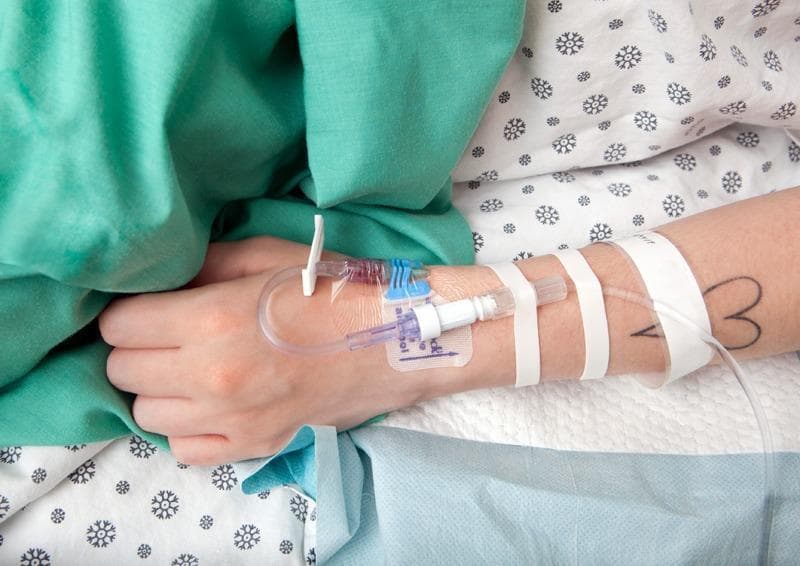 A clinical photograph depicting an intravenous (IV) catheterization on a patient's left forearm in an acute care or obstetric setting. The peripheral IV catheter is inserted and secured with a transparent semi-permeable film dressing over the insertion site, allowing for continuous visual monitoring of the site for signs of phlebitis or infiltration. The IV extension set is further stabilized using three strips of white medical adhesive tape across the forearm to prevent accidental dislodgement. The setup includes a blue slide clamp and a needleless connector capped with a white protective cover. A small amount of dark fluid, consistent with blood flashback, is visible within the clear hub and extension tubing. The patient is positioned on hospital bedding with a snowflake-patterned sheet and a green surgical gown or drape. This procedural image demonstrates standard clinical practice for vascular access and securement in managing complications such as gestational diabetes or preeclampsia.