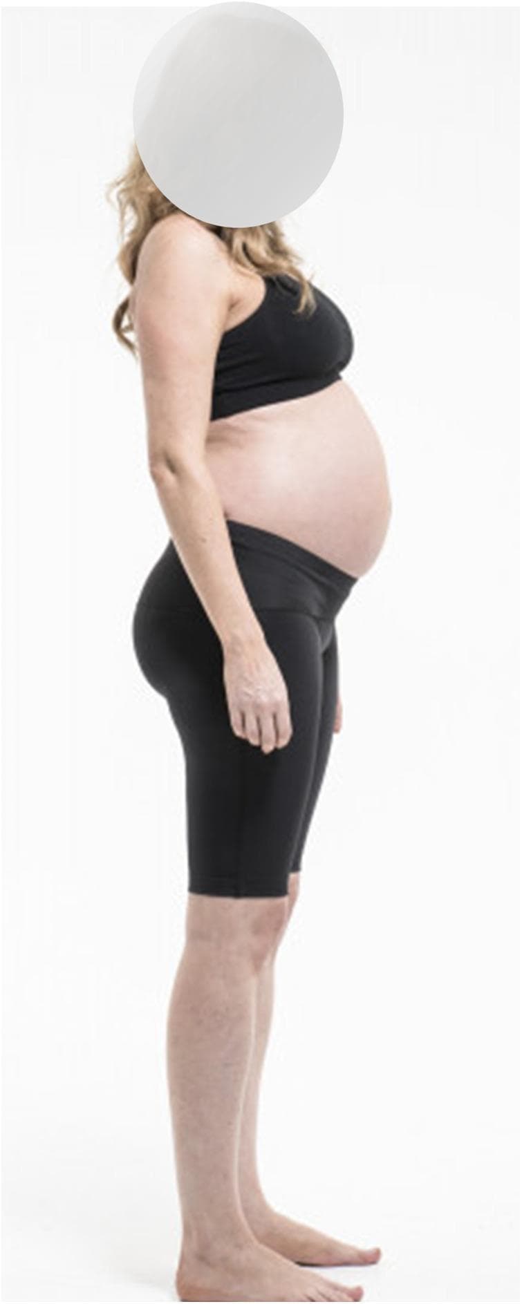 Clinical photograph of a pregnant individual in the third trimester, captured from a lateral profile view to demonstrate anatomical changes and the use of maternity support garments. The image highlights the physiological abdominal protrusion and the compensatory postural adjustments typical of late pregnancy, including an increased lumbar lordosis (swayback) and slightly posterior positioning of the shoulders. The individual is wearing black maternity support shorts (Dynamic Elastomeric Fabric Orthoses) designed with a high waistband that sits beneath the abdominal curve to provide pelvic and lower back stability. This visual serves to illustrate the mechanical management of pregnancy-related pelvic girdle pain (PGPP) and low back pain (LBP) through compression therapy. The educational focus is on maternal health, obstetrics, and physical therapy interventions for musculoskeletal changes during gestation.