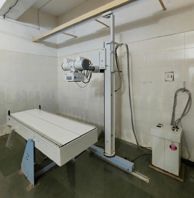 A clinical photograph of a stationary diagnostic X-ray machine within a medical facility. The equipment comprises three primary components: a radiographic examination table, a ceiling-and-floor mounted X-ray tube assembly, and a generator control unit. The examination table is a flat, white, rectangular platform supported by a blue-framed pedestal base. The X-ray source (tube housing and collimator) is mounted on a vertical support column that travels along a floor-mounted rail and a ceiling track, allowing for longitudinal positioning over the patient. Thick insulated high-voltage cables connect the X-ray tube to a floor-standing generator cabinet. The setting features clinical infrastructure including white-tiled walls and a dark, non-porous linoleum floor. This visual serves to illustrate the fundamental hardware configuration used in conventional radiography for skeletal and thoracic diagnostic imaging.