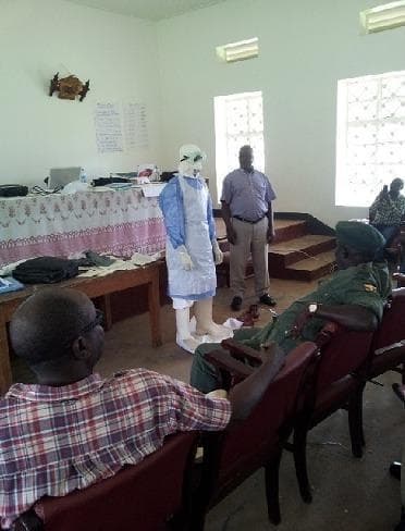 This clinical photograph captures a Personal Protective Equipment (PPE) demonstration during a medical training session focused on Viral Hemorrhagic Fever (VHF) preparedness, specifically for Ebola Virus Disease (EVD). A participant is shown wearing a complete, multi-layered PPE ensemble which includes a white head hood, protective goggles, a light blue long-sleeved isolation gown, a white fluid-resistant apron, white gloves, and high-top white boot covers. The training takes place in a classroom setting with other participants and instructors visible, emphasizing the 'donning and doffing' protocol necessary for infection prevention and control (IPC). The visual illustrates standard barrier nursing precautions and high-level protection required for managing highly infectious pathogens in healthcare or border surveillance contexts. Key educational concepts include contact and droplet precautions, biocontainment procedures, and the role of multi-disciplinary teams in epidemic response.