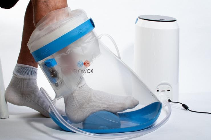 A clinical photograph illustrating the FlowOxβ’ therapeutic device, a specialized medical system used for managing peripheral arterial disease (PAD) and critical limb ischemia. The image displays a patient's lower leg and foot, wearing a white sock, positioned inside a transparent, conical 'boot' chamber. The boot features a blue base and a blue adjustable top band, connected to a translucent upper sleeve that creates an airtight seal around the lower leg. The foot rests on a blue, dome-shaped internal platform designed to prevent pressure points. A clear pneumatic tube connects the boot's base to a white, cylindrical control unit that generates pulsed intermittent negative pressure (typically -40 mmHg). This therapy is designed to enhance blood flow and microcirculation in the lower extremities for patients with reduced ankle-brachial pressure index (ABPI) or claudication. The device is shown in a clinical or home-use setting, emphasizing its role as a non-invasive vascular intervention for limb salvage and wound care.