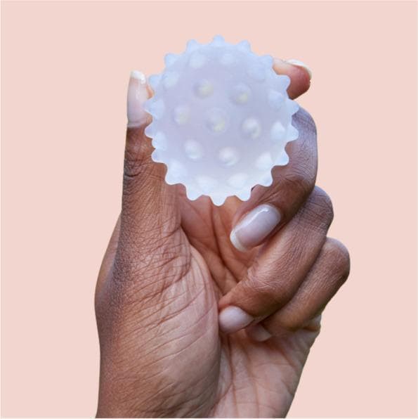 A clinical photograph of a Lactamo device, a specialized lactation aid designed for maternal health and breastfeeding support. The device is a small, handheld sphere, approximately 5 cm in diameter, constructed from a translucent, frosted medical-grade material. Its surface is characterized by multiple rounded, hollow protrusions or nodes distributed evenly across the sphere to provide varying levels of compression and tactile stimulation. The device is designed to be filled with a thermochromic gel, allowing for the delivery of hot or cold therapy in conjunction with breast massage and compression. It is used to address breastfeeding challenges such as engorgement, blocked ducts, and mastitis by facilitating milk flow through movement toward the nipple or axilla. The photograph shows the device held between fingers to demonstrate its scale and ergonomic design for self-application by postpartum patients.
