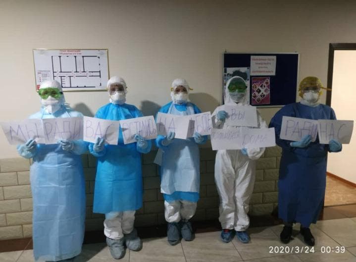 A clinical photograph depicting five healthcare workers in a medical facility during the COVID-19 pandemic, illustrating standard and enhanced personal protective equipment (PPE) protocols. The workers are wearing a variety of PPE configurations, including disposable fluid-resistant gowns in light and dark blue, white full-body coveralls (tyvek suits), surgical caps, and hoods. Essential infection control components visible include N95 or surgical face masks, protective goggles, face shields, and blue nitrile gloves. The photograph is set in a hospital corridor, featuring a wall-mounted evacuation floor plan and a bulletin board in the background. The image serves as a visual record of public health frontline response, highlighting the variability in PPE styles and the utilization of contact and airborne precautions in a clinical setting during a global respiratory viral outbreak.